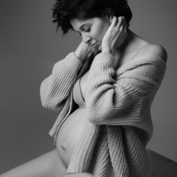 Aleksandra Lauda Fotografia - Czarno-białe, portretowe ujęcie kobiety w ciąży, siedzącej bokiem, z rękami na głowie, w rozpiętym swetrze, z delikatnym uśmiechem i wzrokiem skierowanym w dół.