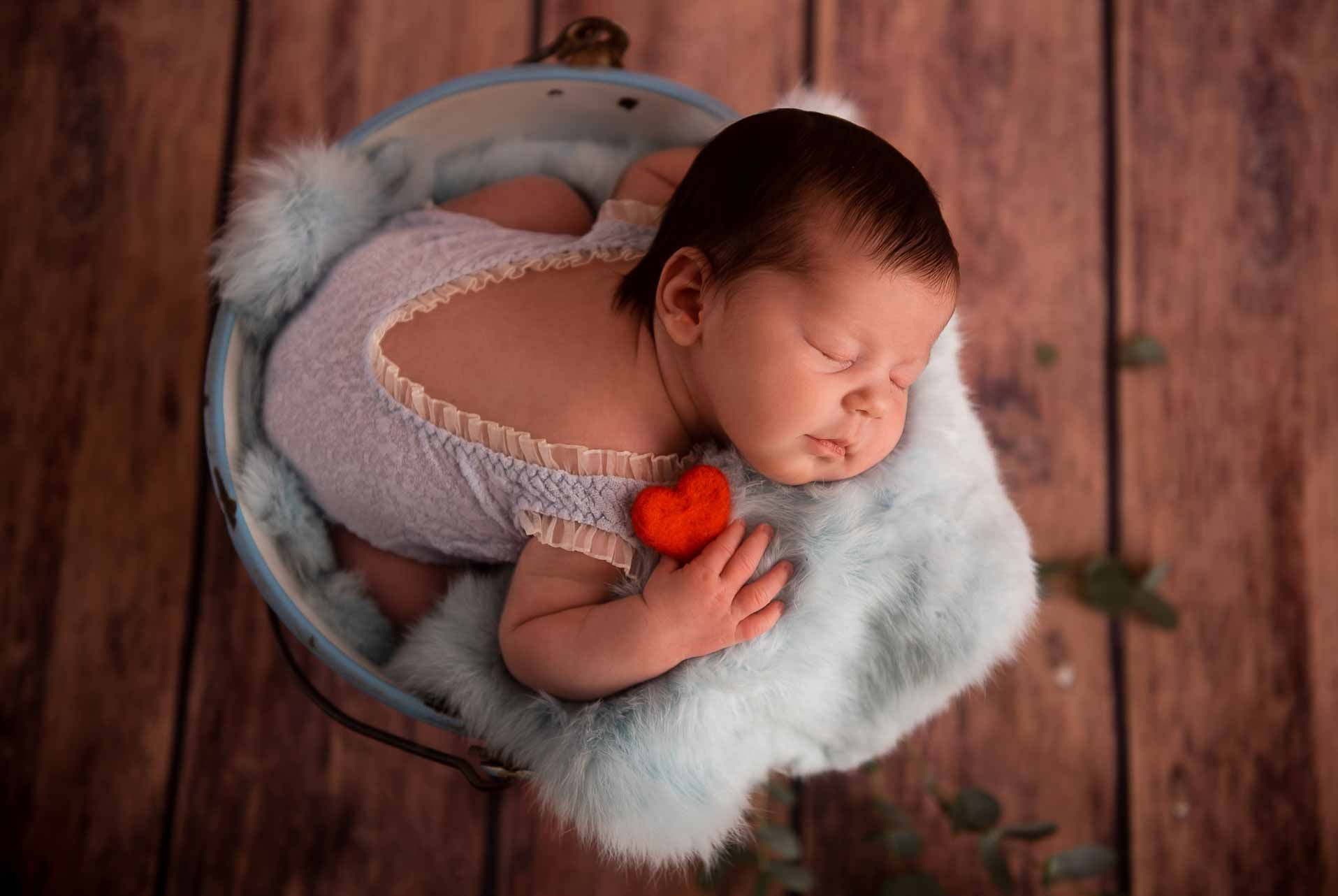 Śpiący noworodek w błękitnym ubranku, ułożony w metalowym wiaderku wyłożonym futerkiem, z małym czerwonym serduszkiem w dłoni.