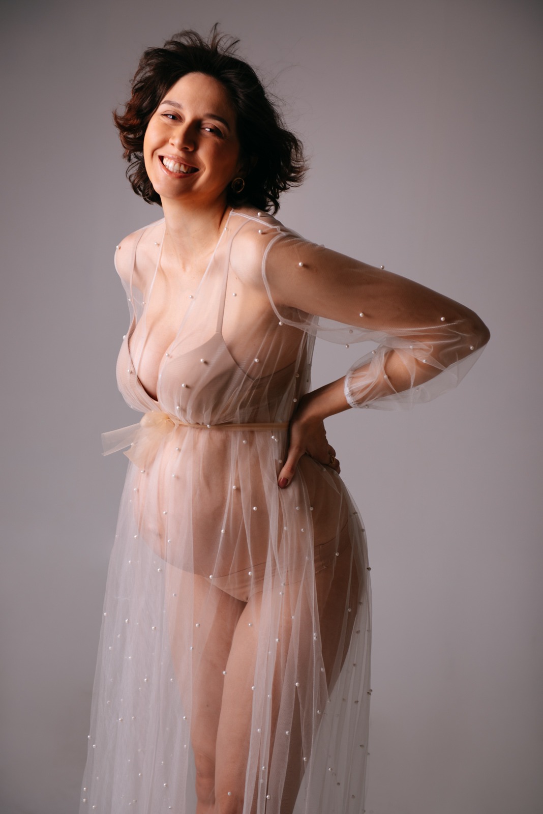 Uśmiechnięta kobieta w ciąży, pozująca w przezroczystej, tiulowej sukni zdobionej perłami, z ręką na biodrze, na jasnoszarym tle.