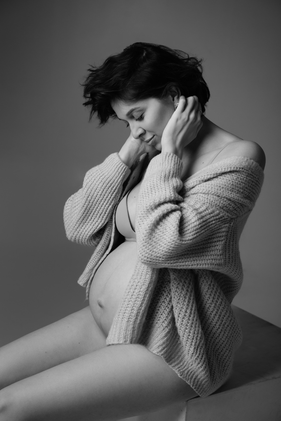 Czarno-białe, portretowe ujęcie kobiety w ciąży, siedzącej bokiem, z rękami na głowie, w rozpiętym swetrze, z delikatnym uśmiechem i wzrokiem skierowanym w dół.