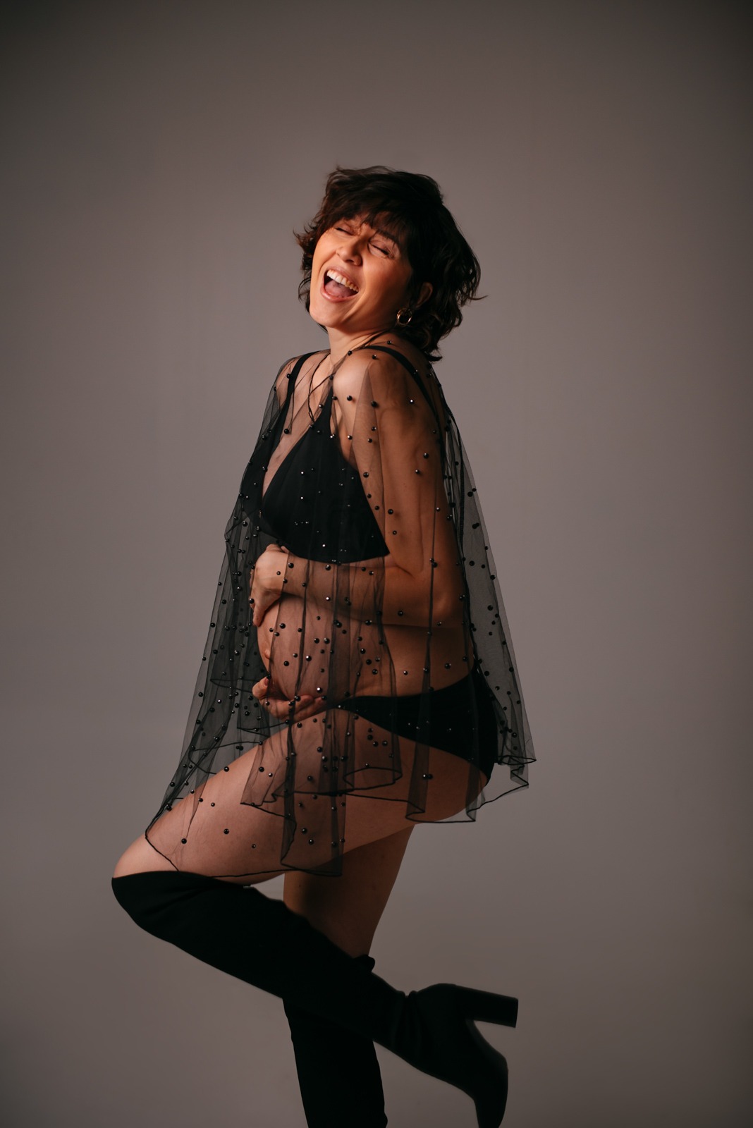 Kobieta w ciąży, ubrana w czarny strój i przezroczystą narzutkę w kropki, pozuje z uniesioną nogą i szerokim uśmiechem na szarym tle.