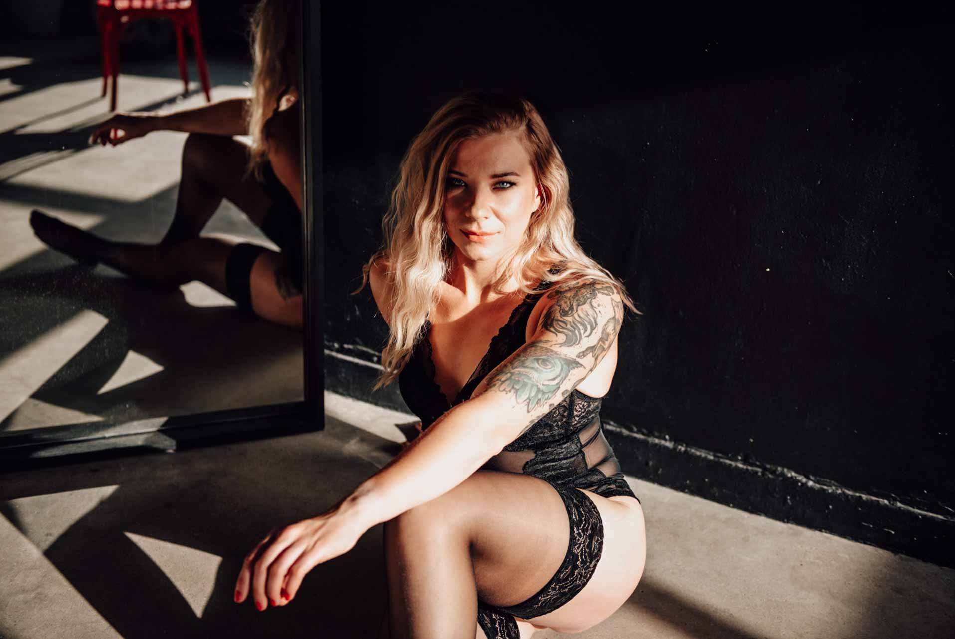Kobieta w czarnej bieliźnie, pozująca w studio z ciemnym tłem i lustrem, podkreślona promieniami światła tworzącymi geometryczne cienie na podłodze.