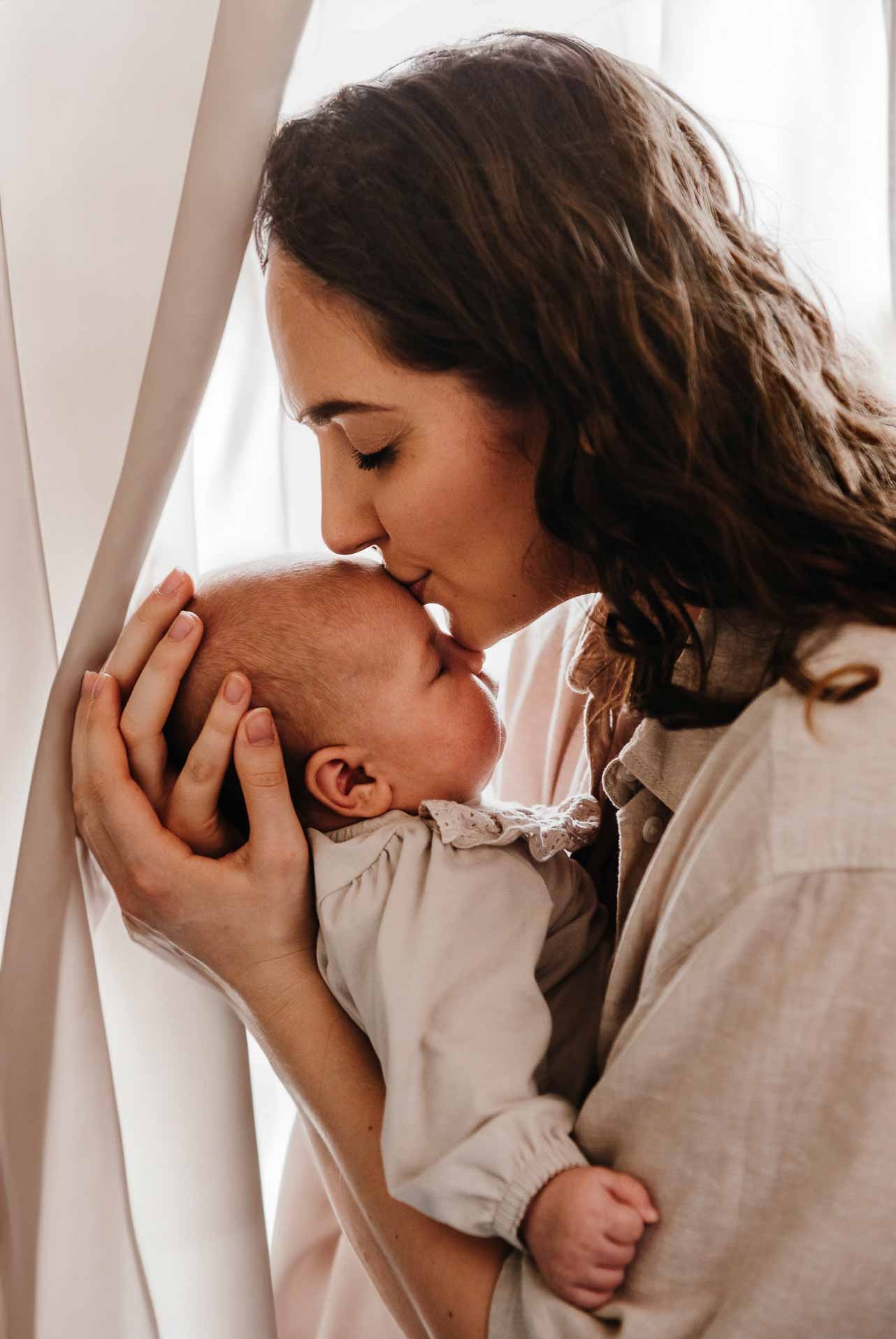Mama z zamkniętymi oczami całuje w główkę śpiące niemowlę, trzymając je w ramionach przy oknie z jasną zasłoną.