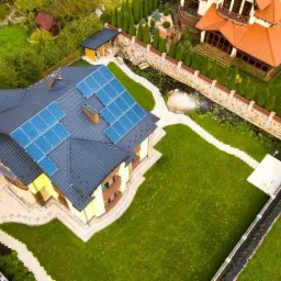 Widok z lotu ptaka na dom z panelami słonecznymi na dachu, otoczony zielonym trawnikiem i ozdobnym brukiem.