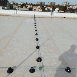 ELE-MAX sp z o.o. - System odgromowy z czarnymi uchwytami na płaskim dachu budynku wielorodzinnego, widoczny cień osoby fotografującej.