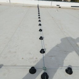 ELE-MAX sp z o.o. - Instalacja odgromowa z czarnymi uchwytami na płaskim dachu budynku, widoczne metalowe linki i złącza krzyżowe, tło stanowią budynki mieszkalne i drzewa.