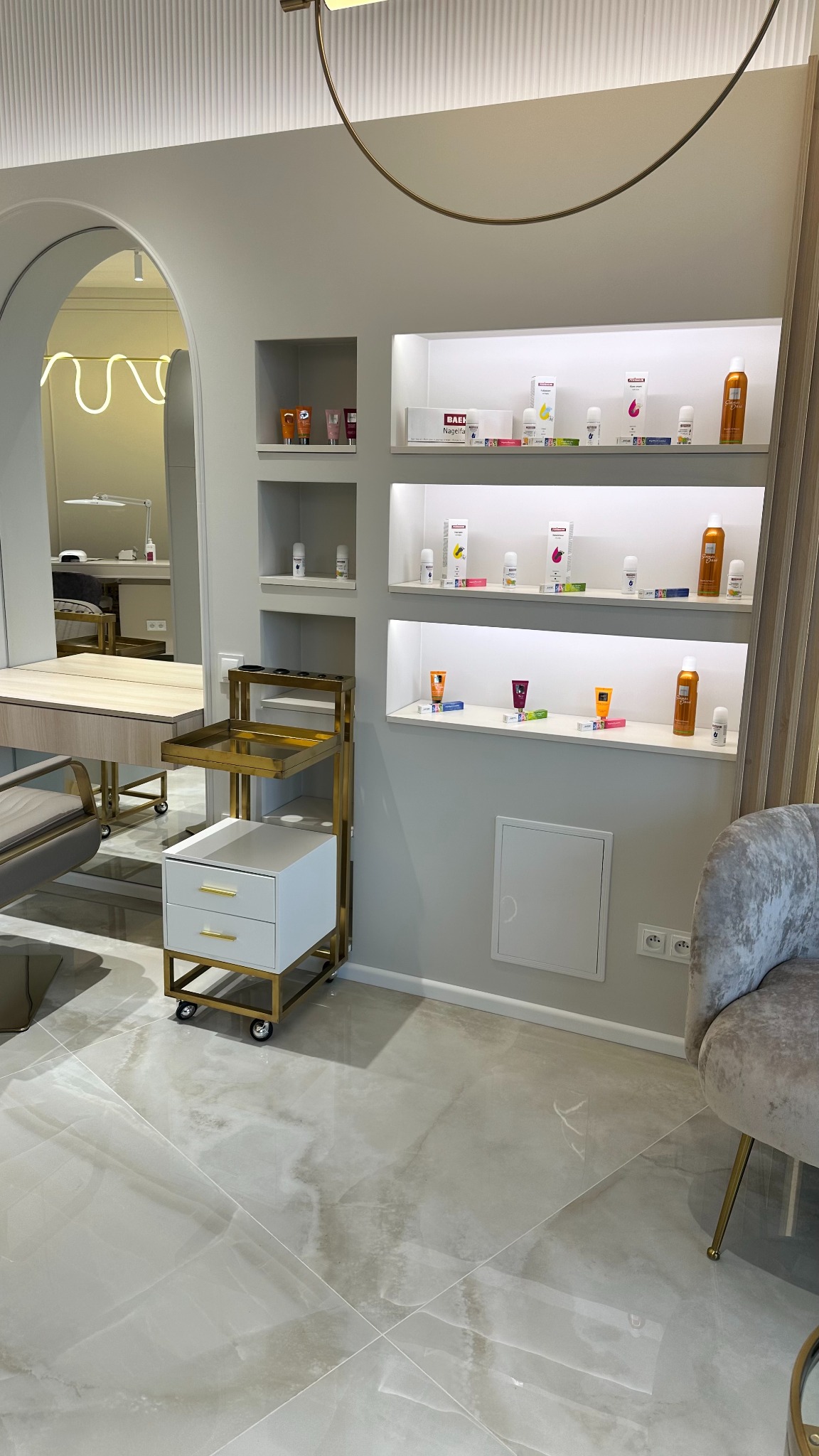 Jasne wnętrze salonu kosmetycznego z regałami pełnymi kosmetyków, złotym wózkiem z szufladami i lustrem w kształcie łuku.