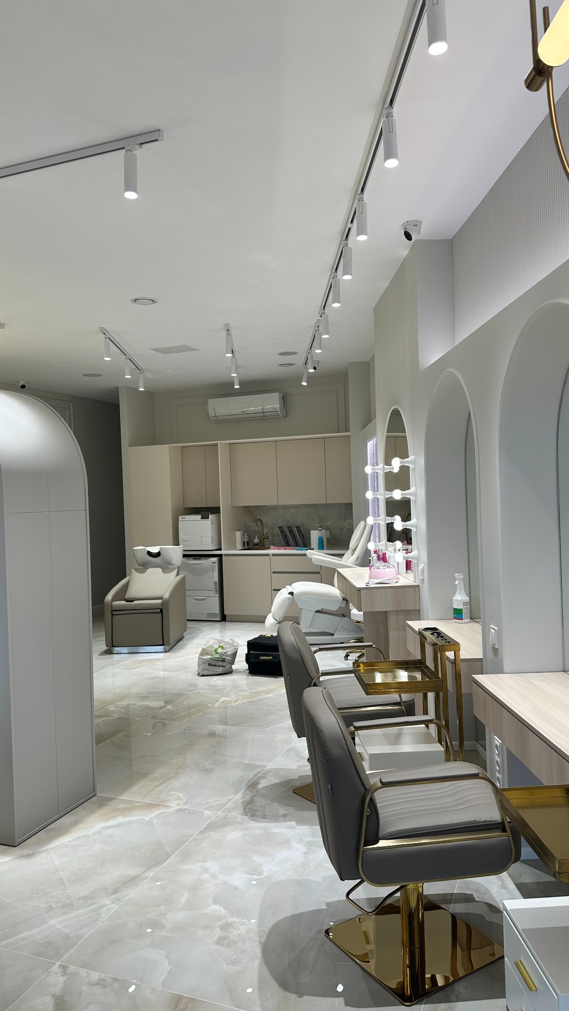Wnętrze salonu kosmetycznego z szarymi fotelami na złotych podstawach, oświetleniem punktowym na suficie, stanowiskami do manicure z lustrami podświetlanymi żarówkami i widocznym systemem monitoringu.