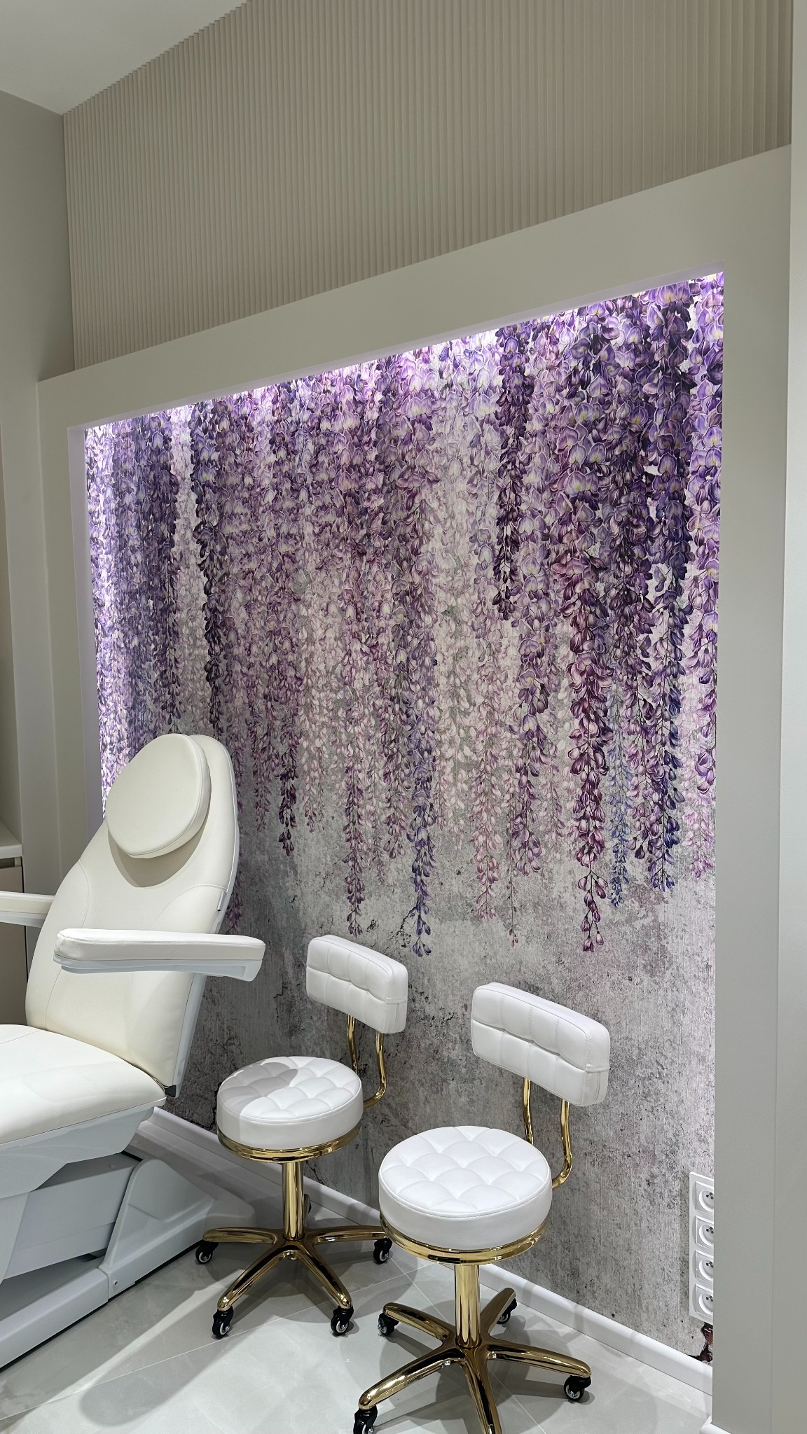 Białe, skórzane krzesła kosmetyczne na złotych podstawach z kółkami, ustawione przed ścianą z motywem kwitnącej glicynii w salonie kosmetycznym. W tle widoczny fotel kosmetyczny.