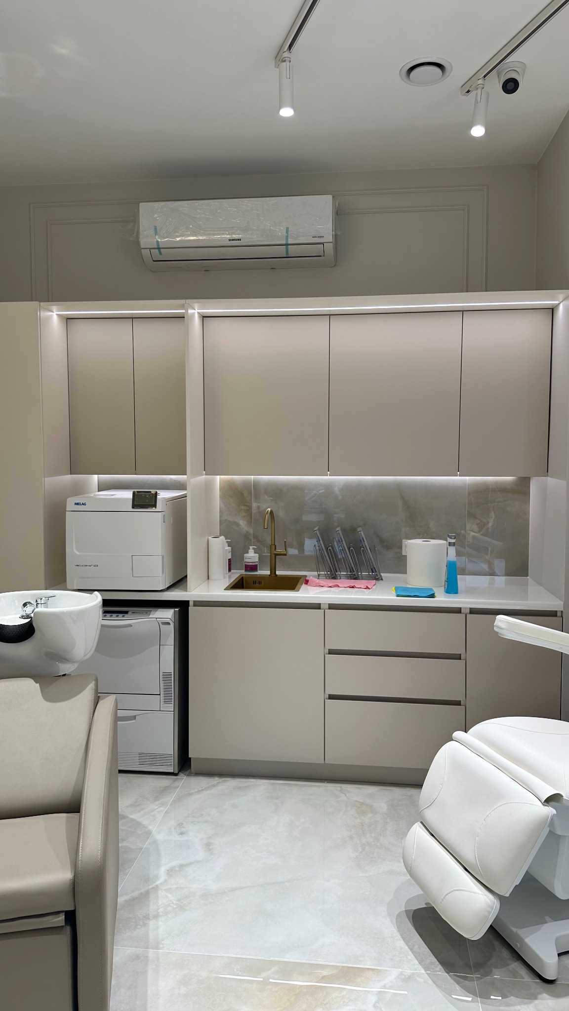 Kremowe szafki w gabinecie dentystycznym z marmurowym blatem, złotą baterią, sterylizatorem MELAG i unit dentystyczny z białym fotelem.