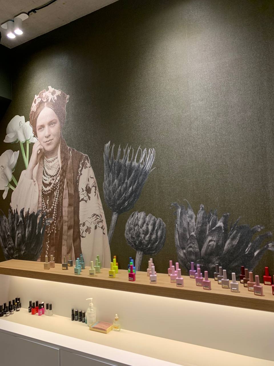 Ściana salonu kosmetycznego z fototapetą przedstawiającą portret kobiety w stroju ludowym i motywy roślinne; na półkach prezentacja kolorowych lakierów do paznokci, żeli antybakteryjnych i innych...