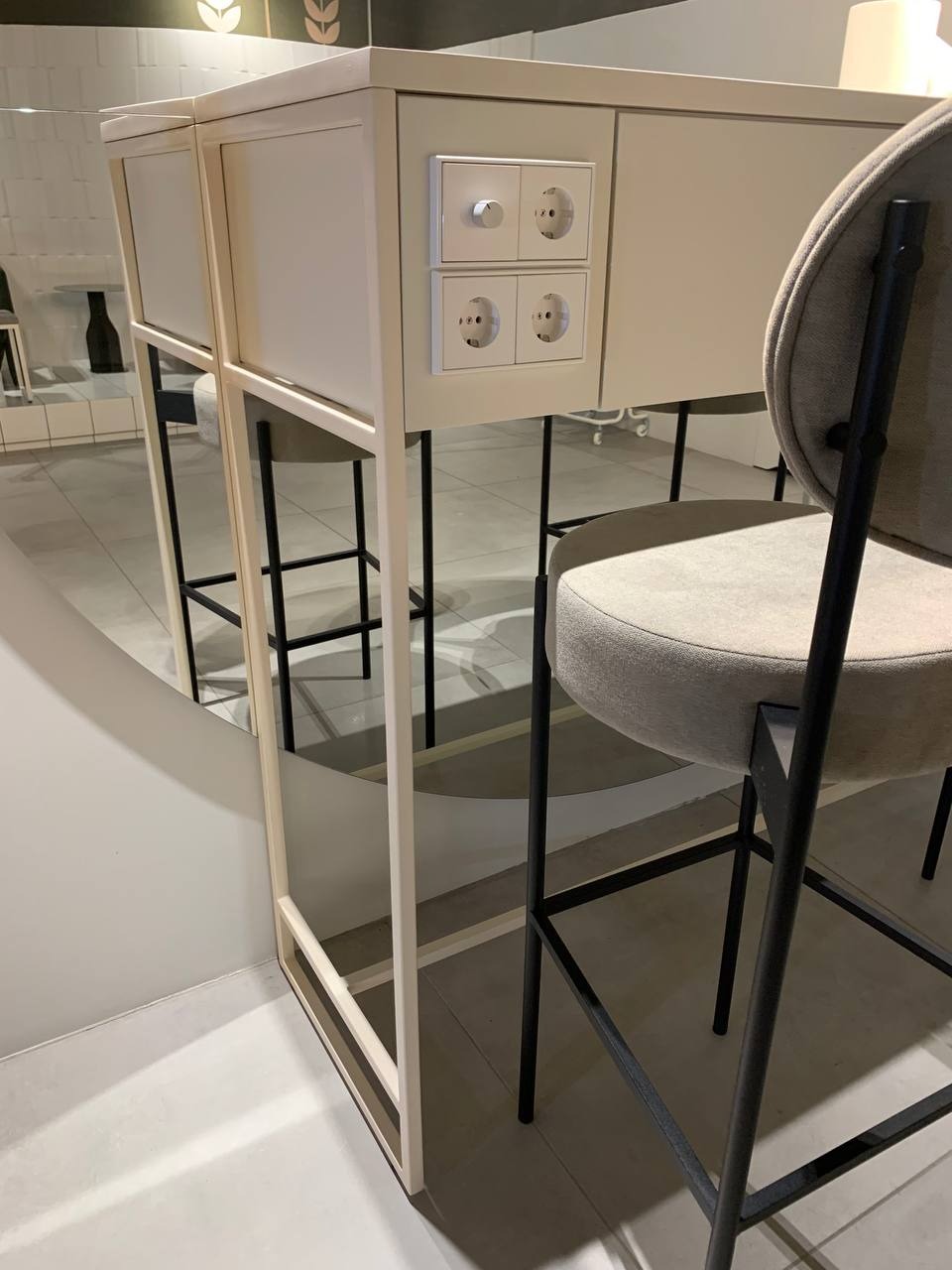 Dwa gniazdka elektryczne i ściemniacz zamontowane w blacie beżowej, minimalistycznej wyspy kuchennej z krzesłem barowym, widoczne w lustrzanym odbiciu.