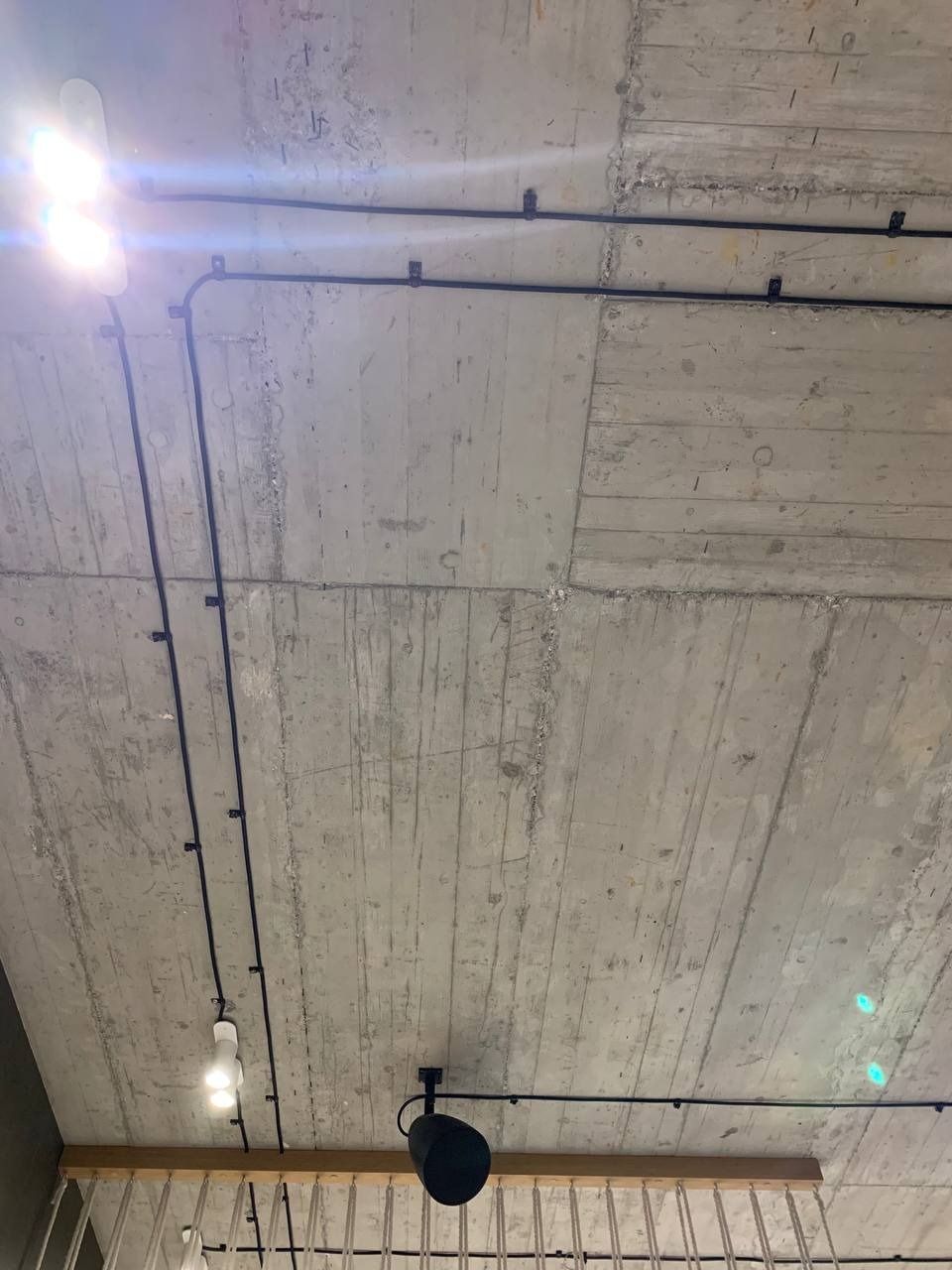 Betonowy sufit z widocznymi czarnymi kablami elektrycznymi, zamocowanymi do powierzchni za pomocą uchwytów, z zamontowanym oświetleniem punktowym i lampą wiszącą.