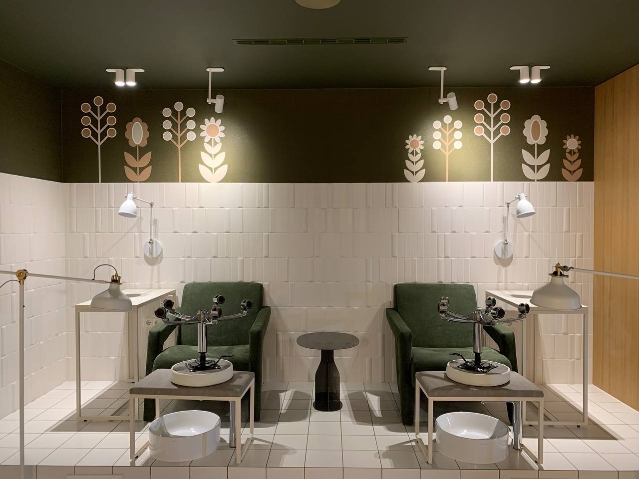 Nowoczesne stanowisko do zabiegów kosmetycznych z zielonymi fotelami, białymi umywalkami i lampami, dekorowane motywami roślinnymi na ścianie w odcieniach brązu i beżu.