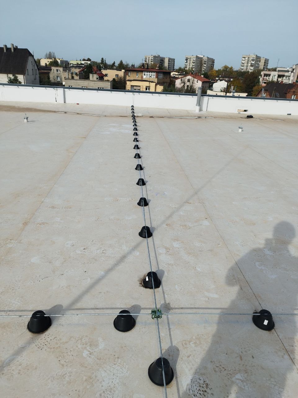 System odgromowy z czarnymi uchwytami na płaskim dachu budynku wielorodzinnego, widoczny cień osoby fotografującej.