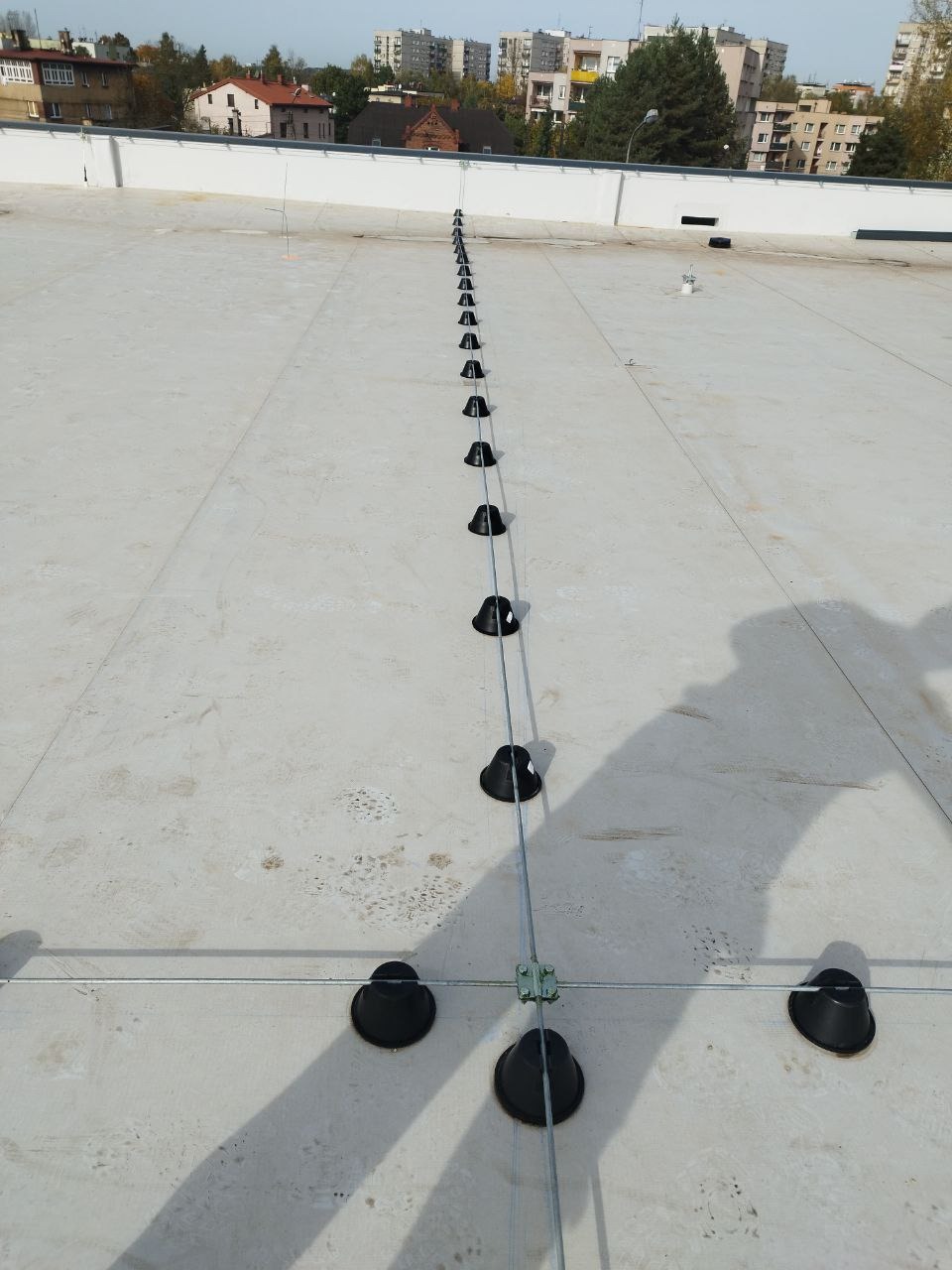 Instalacja odgromowa z czarnymi uchwytami na płaskim dachu budynku, widoczne metalowe linki i złącza krzyżowe, tło stanowią budynki mieszkalne i drzewa.