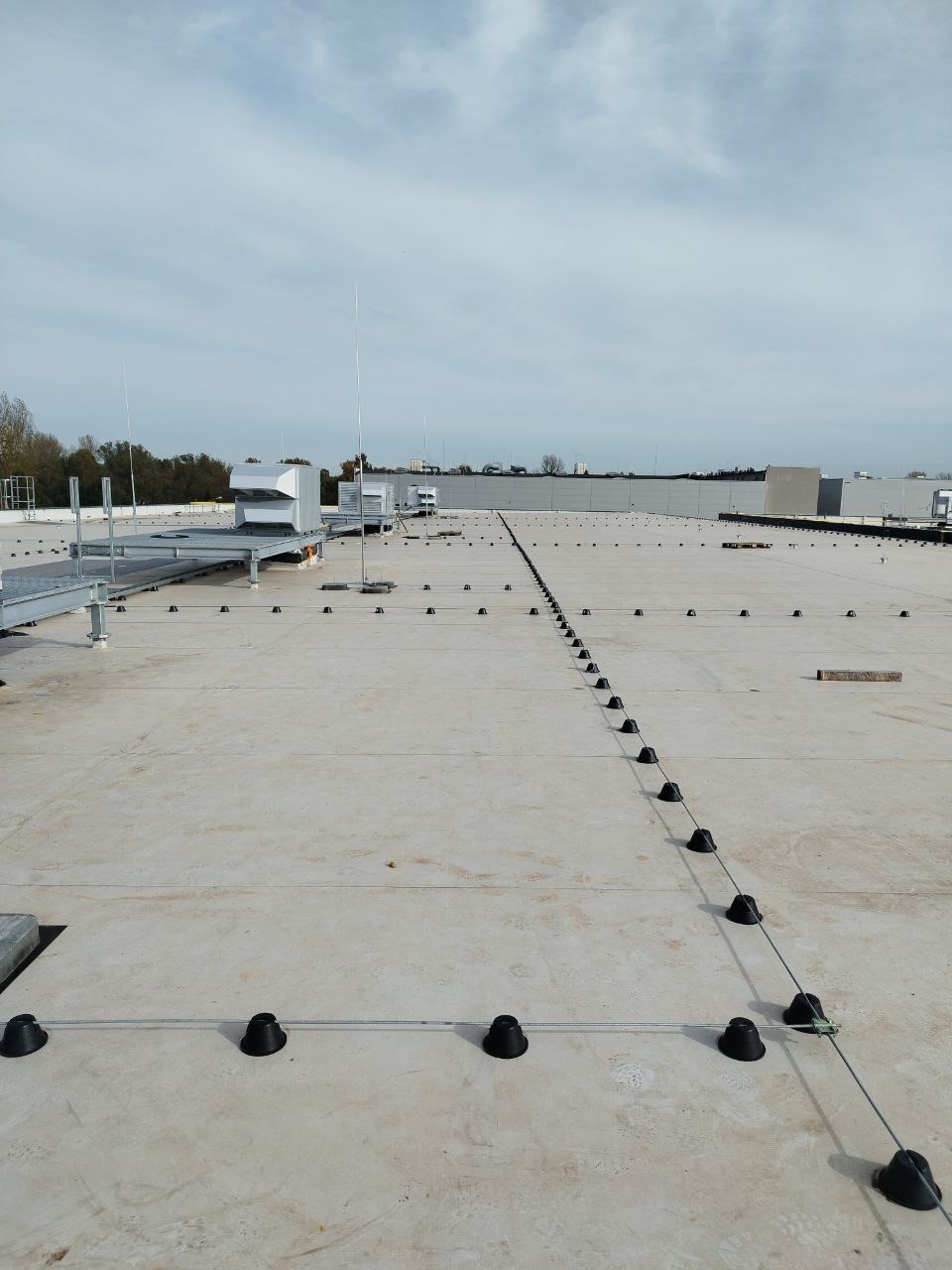 Instalacja odgromowa na płaskim dachu budynku przemysłowego, widoczne przewody i uchwyty mocujące, w tle urządzenia wentylacyjne i maszty.