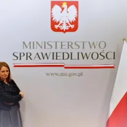 Kobieta z założonymi rękami na tle ściany z godłem i napisem Ministerstwo Sprawiedliwości oraz trzema polskimi flagami.