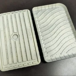 Dwie metalowe formy odlewnicze o różnych wzorach, z licznymi otworami, leżące na ciemnej powierzchni. Detal obróbki CNC.