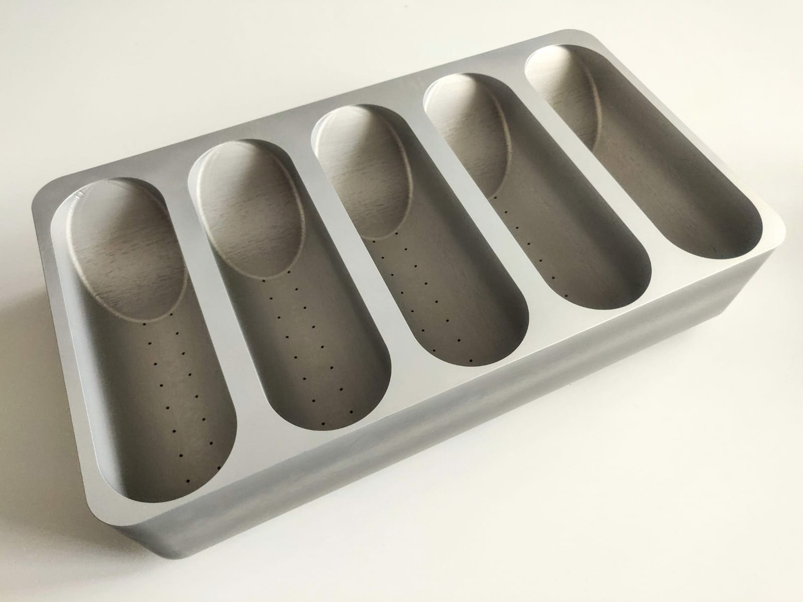 Szara, metalowa forma z pięcioma podłużnymi wgłębieniami z otworami, ustawiona ukośnie na białym tle. Precyzyjna obróbka CNC.