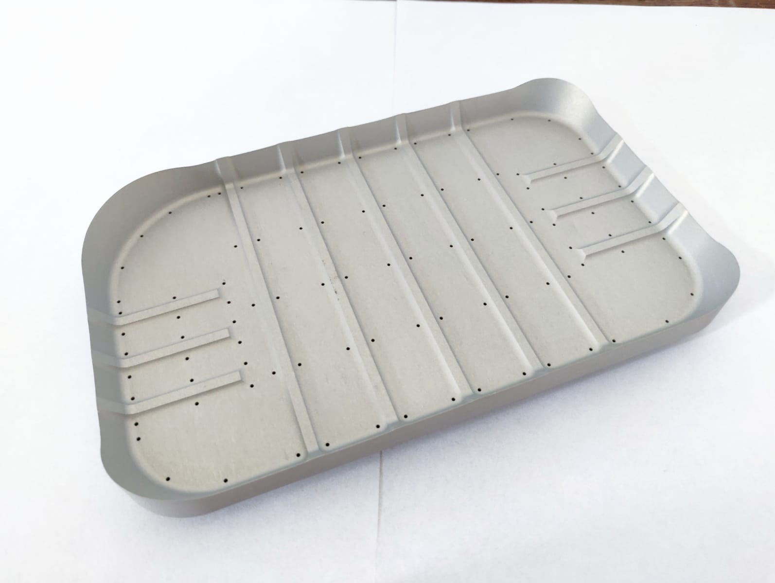Szara, prostokątna forma z metalu z otworami i rowkami, leżąca na białym tle, ukazująca precyzję wykonania i potencjalne zastosowanie w przemyśle.