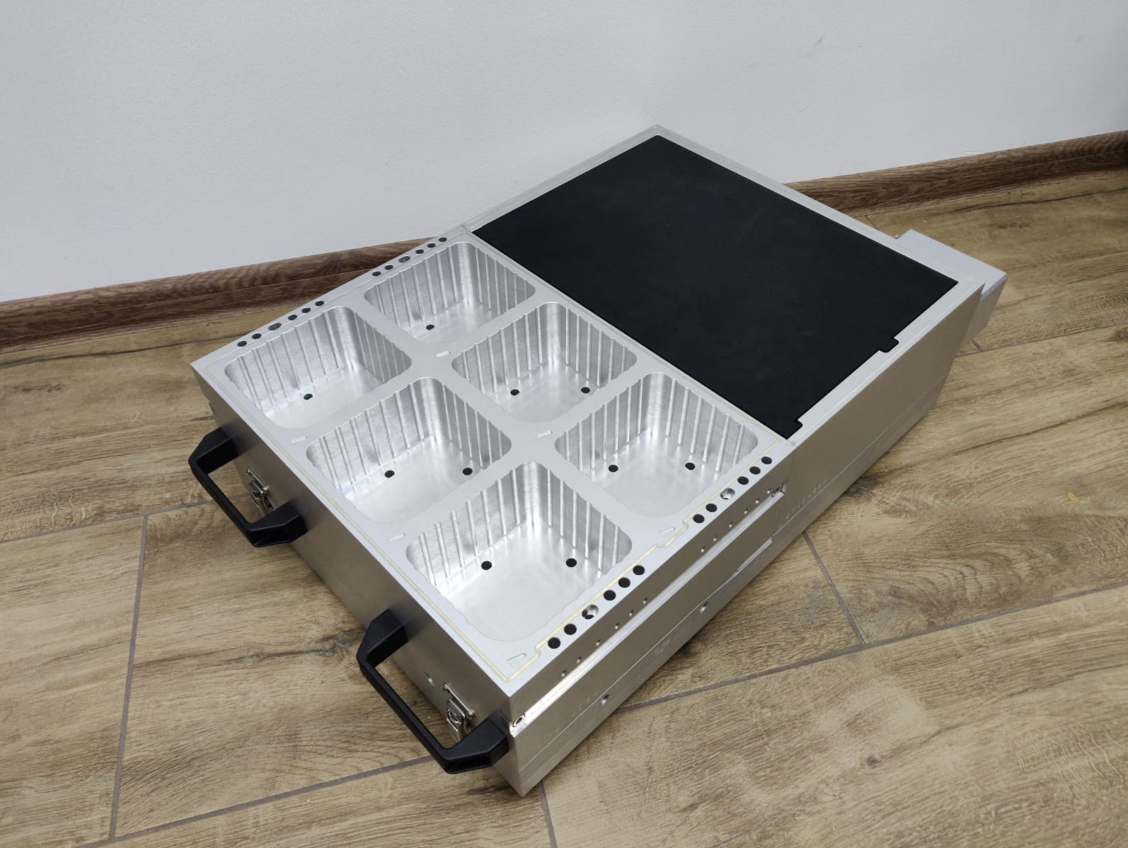 Srebrna, aluminiowa skrzynka z sześcioma przegródkami i czarnym panelem, leżąca na podłodze z paneli. Widoczne uchwyty transportowe i precyzyjne wykonanie detali.
