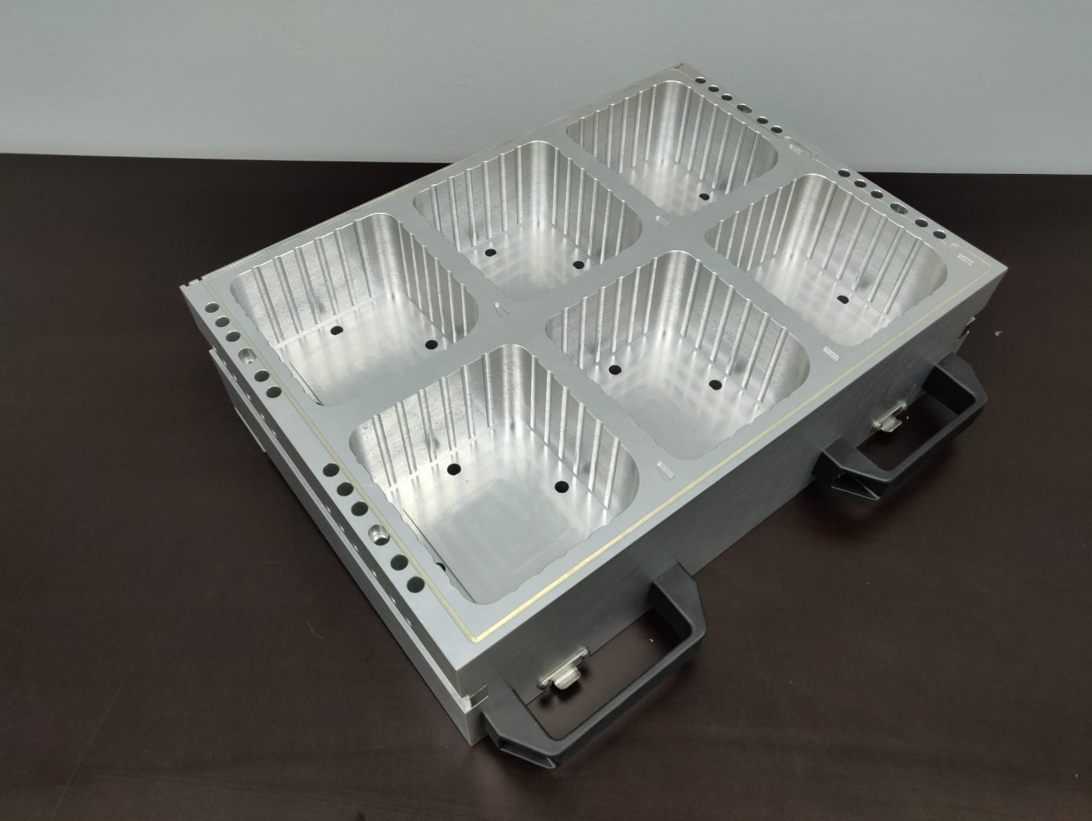 Precyzyjnie obrobiony, srebrny, aluminiowy element z sześcioma wgłębieniami, widoczne otwory i żłobienia, na ciemnym blacie, z uchwytami po bokach.