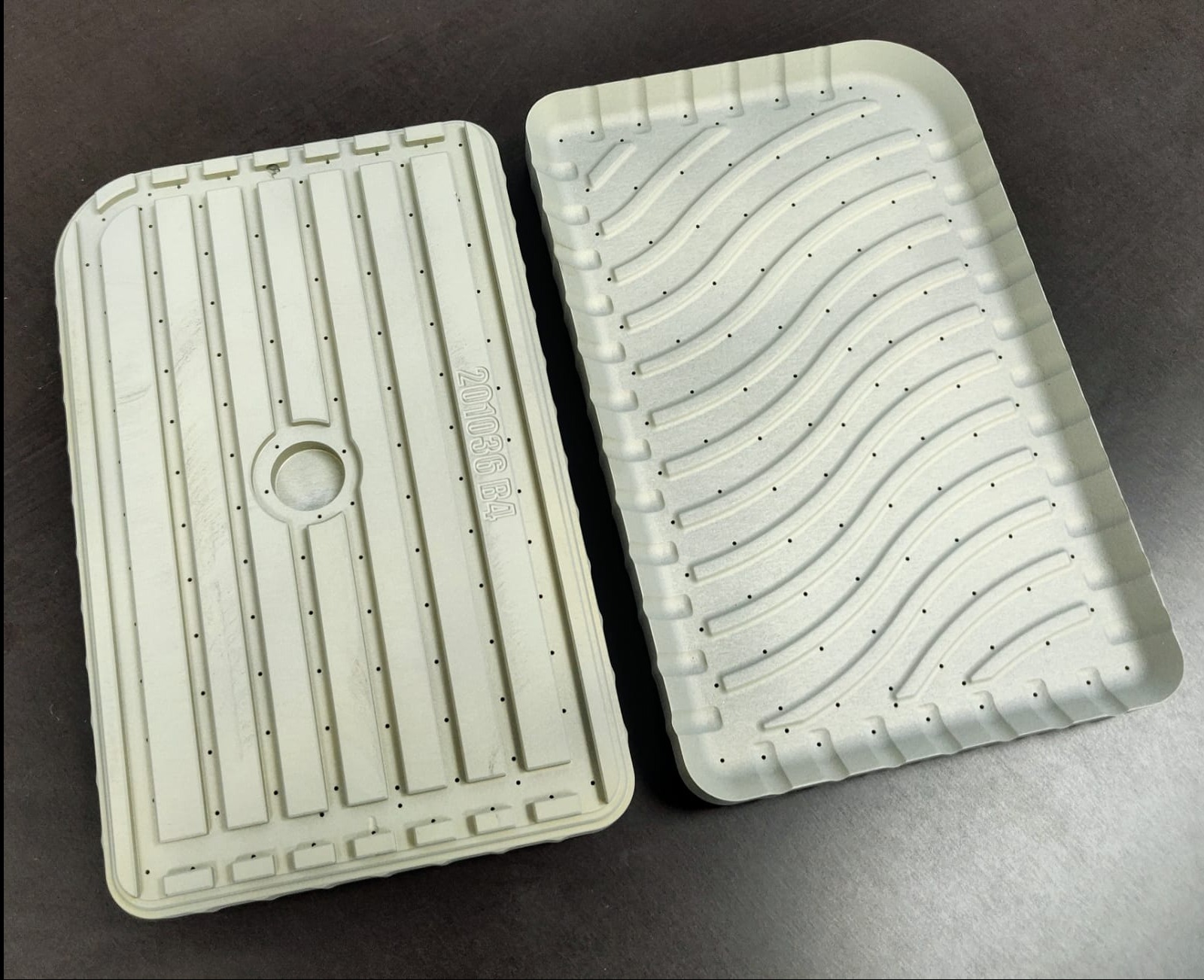 Dwie metalowe formy odlewnicze o różnych wzorach, z licznymi otworami, leżące na ciemnej powierzchni. Detal obróbki CNC.