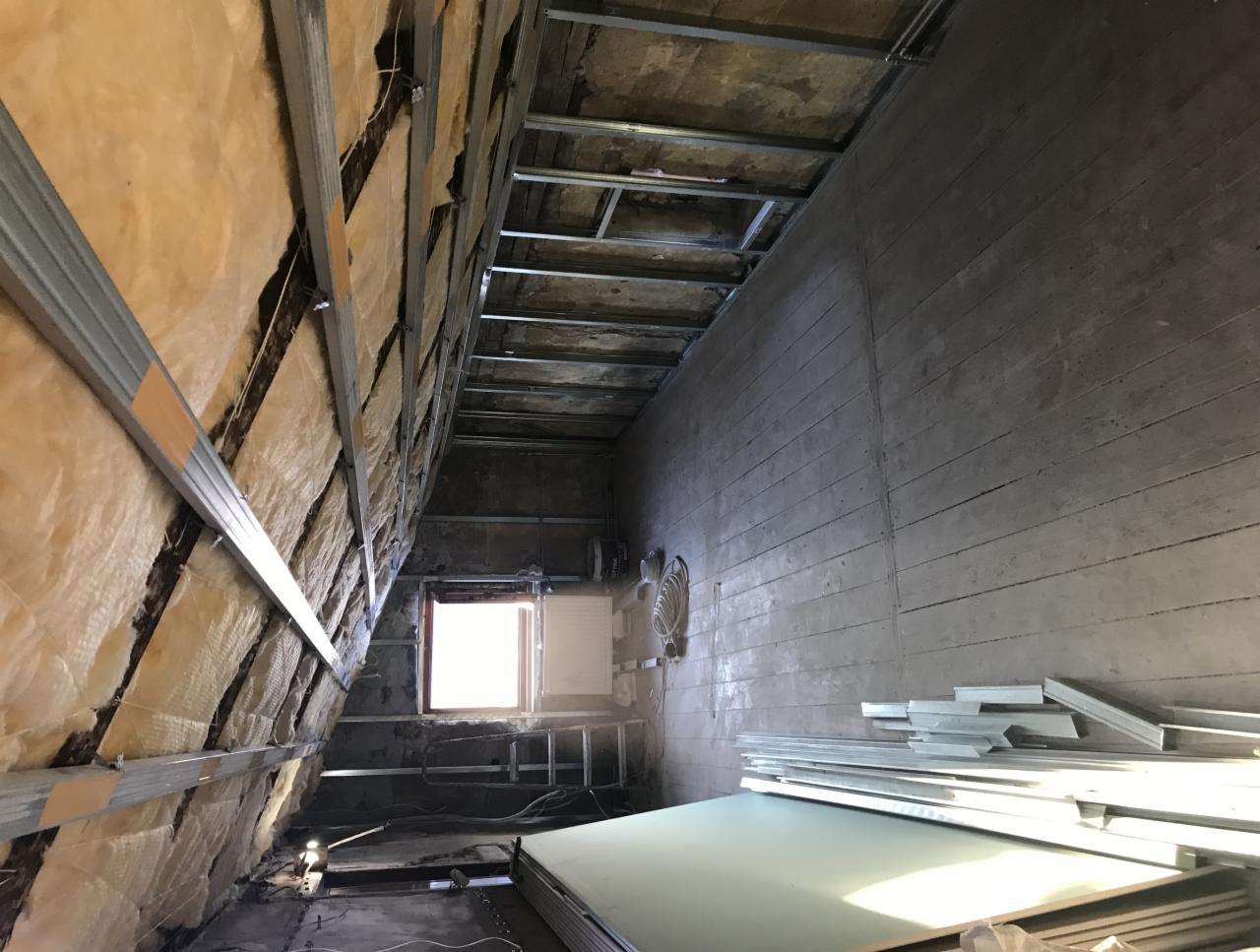 Adaptacja poddasza: widok na ścianę z izolacją z wełny mineralnej, metalowy stelaż pod płyty gipsowo-kartonowe oraz stos płyt na podłodze, małe okno w tle.