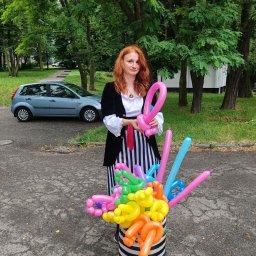 Agencja Usługowo - Artystyczna Kris Krzysztof Kończyk - Kobieta z rudymi włosami w kostiumie pirata trzyma balonowe zwierzątko, obok wiadro z kolorowymi balonami modelowanymi, w tle drzewa i zaparkowany niebieski samochód.