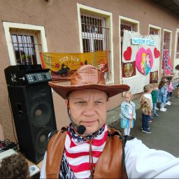 Agencja Usługowo - Artystyczna Kris Krzysztof Kończyk - Mężczyzna w stroju kowbojskim z mikrofonem nagłownym, na tle budynku z dekoracjami i grupką dzieci, obok sprzęt nagłośnieniowy.