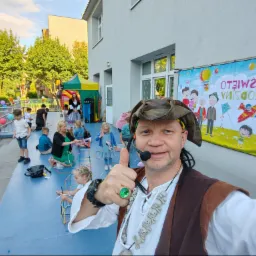 Animator w stroju pirata z mikrofonem, pokazuje kciuk w górę na tle dzieci budujących konstrukcje z rurek i baneru reklamowego z napisem 'Święto Rodziny' podczas imprezy plenerowej.
