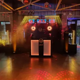 Scena z oświetleniem eventowym: reflektory, kule dyskotekowe, dymiarka. Stanowisko DJ'a z logo. Parkiet oświetlony czerwonymi punktami. Atmosfera imprezy.