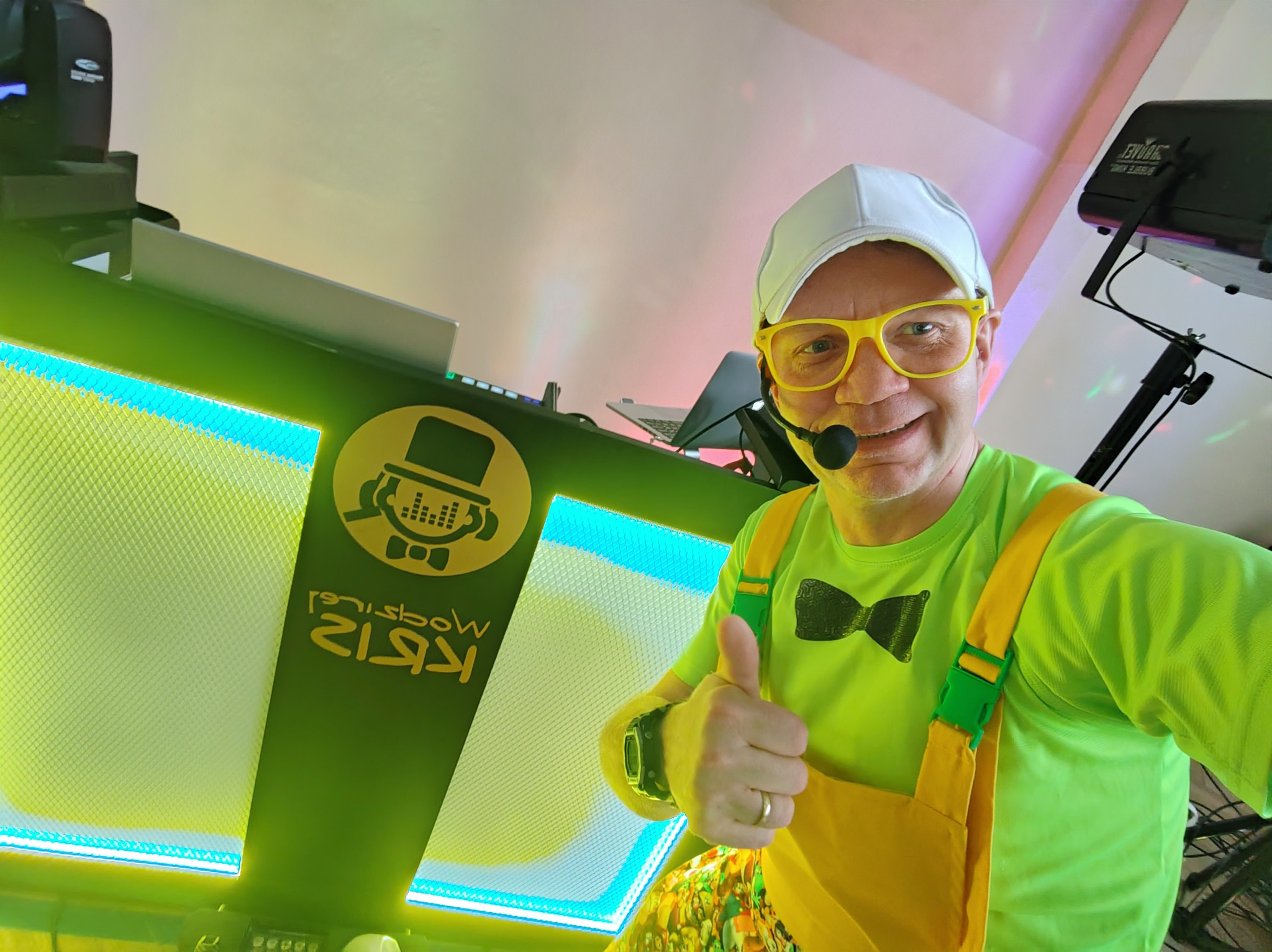 Uśmiechnięty mężczyzna w kolorowym stroju z mikrofonem, żółtych okularach i czapce bejsbolowej, stojący za podświetlanym stanowiskiem DJ-a z logo i sprzętem nagłośnieniowym, kciuk w górę.
