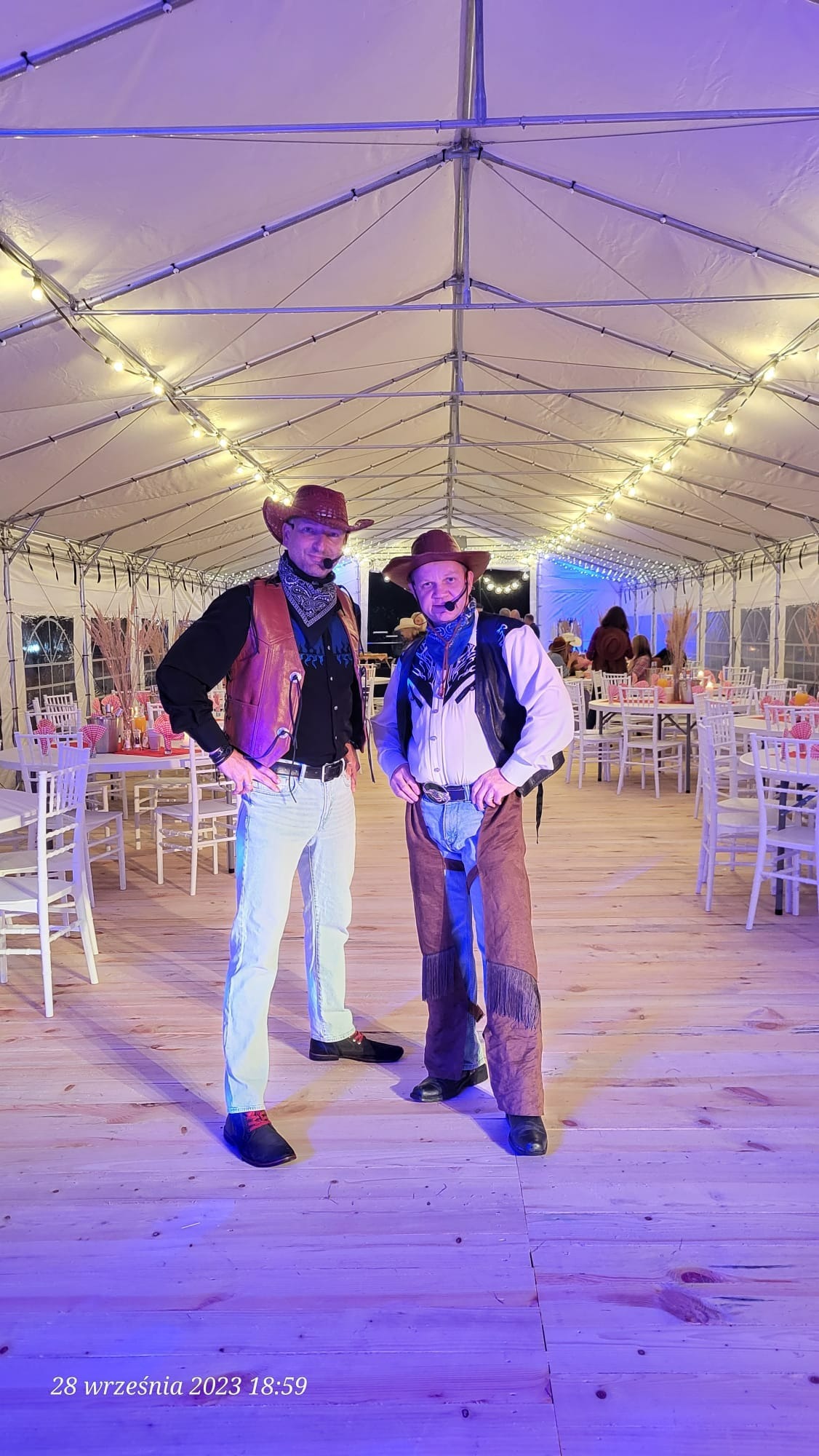 Dwóch mężczyzn w strojach kowbojskich, z mikrofonami nagłownymi, pozuje na drewnianym parkiecie w namiocie z oświetleniem, przygotowanym na imprezę.