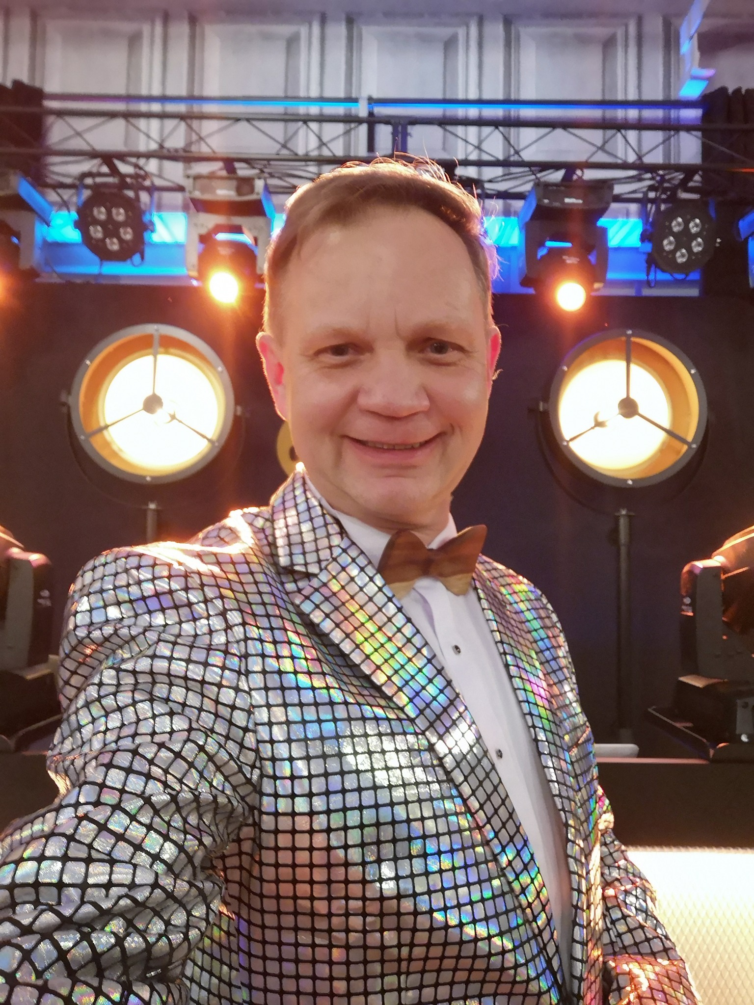 Portret uśmiechniętego mężczyzny w błyszczącej marynarce i drewnianej muszce na tle sceny z reflektorami