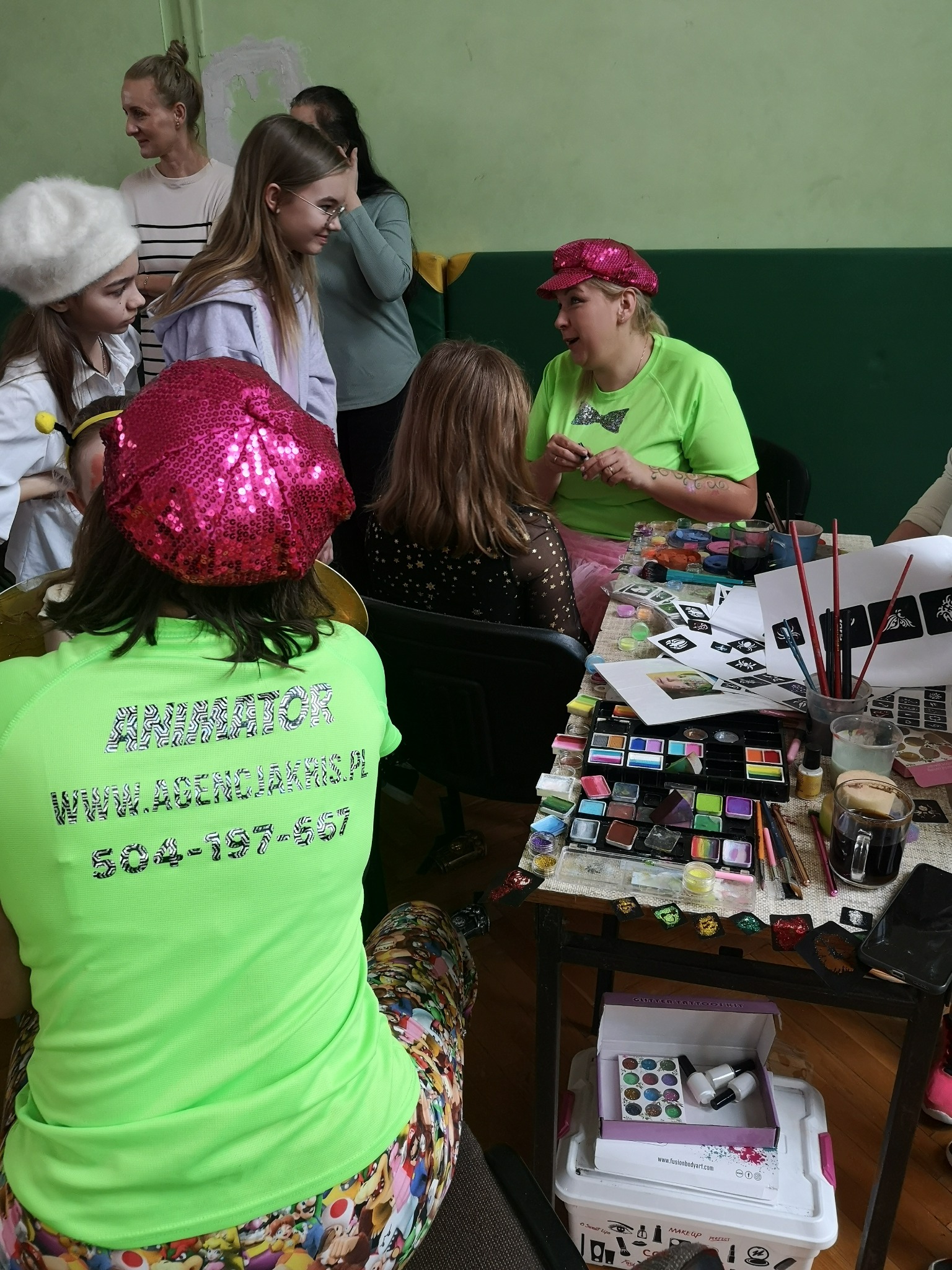 Kobieta w zielonej koszulce z napisem 'ANIMATOR' i różowej cekinowej czapce rozmawia z grupą dzieci i dorosłych, na stole rozłożone farby do malowania twarzy, pędzle i szablony.
