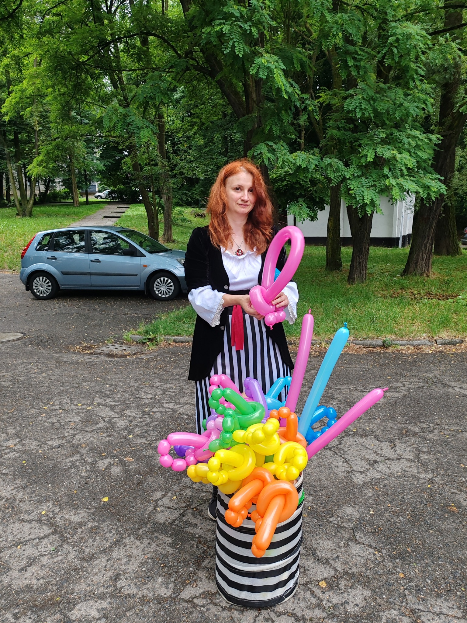 Kobieta z rudymi włosami w kostiumie pirata trzyma balonowe zwierzątko, obok wiadro z kolorowymi balonami modelowanymi, w tle drzewa i zaparkowany niebieski samochód.