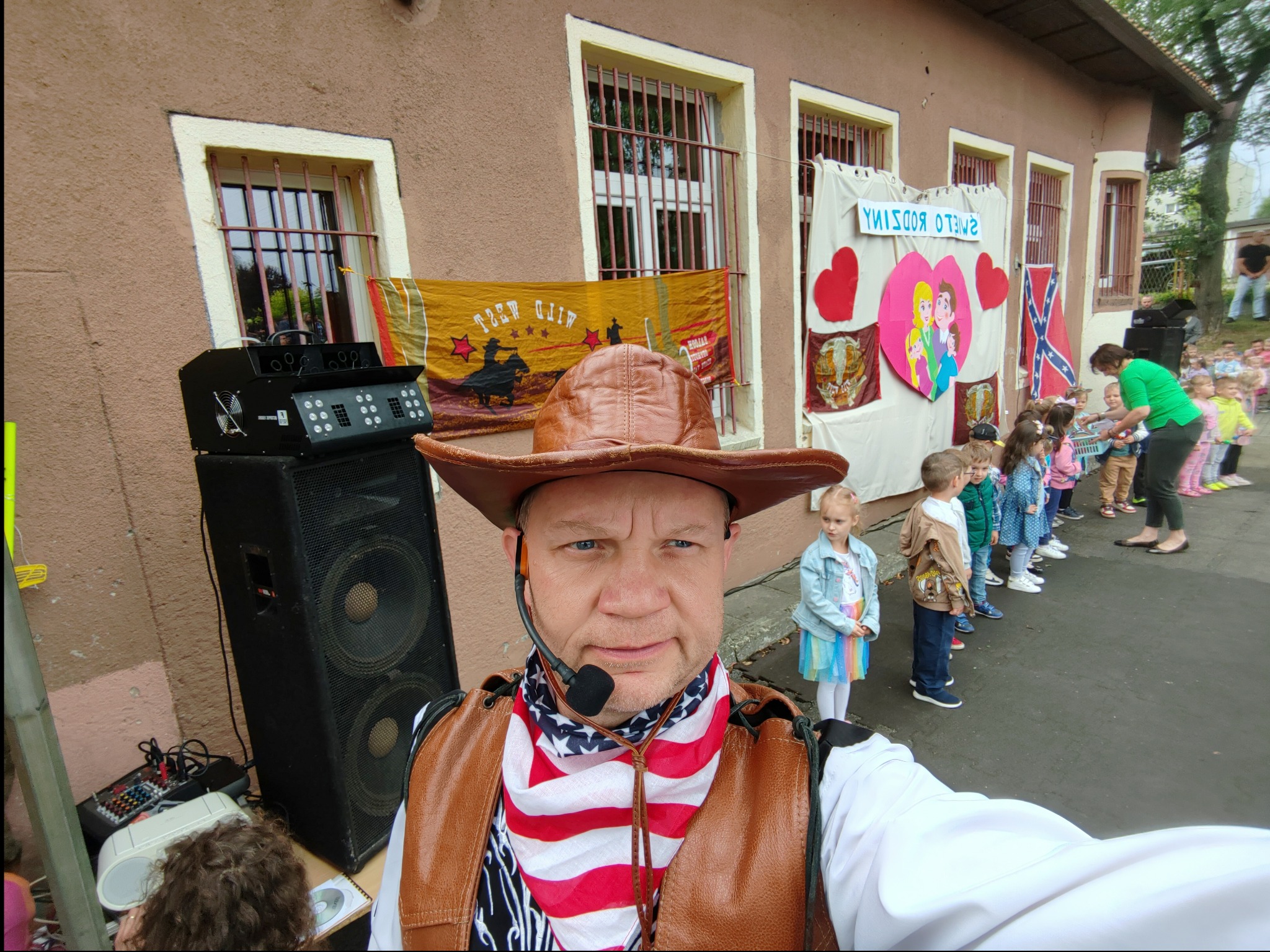 Mężczyzna w stroju kowbojskim z mikrofonem nagłownym, na tle budynku z dekoracjami i grupką dzieci, obok sprzęt nagłośnieniowy.