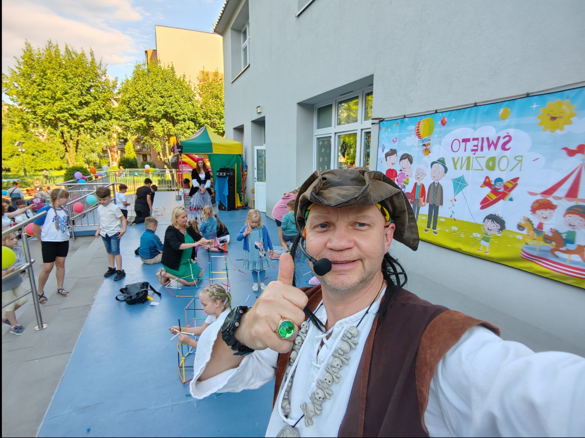 Animator w stroju pirata z mikrofonem, pokazuje kciuk w górę na tle dzieci budujących konstrukcje z rurek i baneru reklamowego z napisem 'Święto Rodziny' podczas imprezy plenerowej.