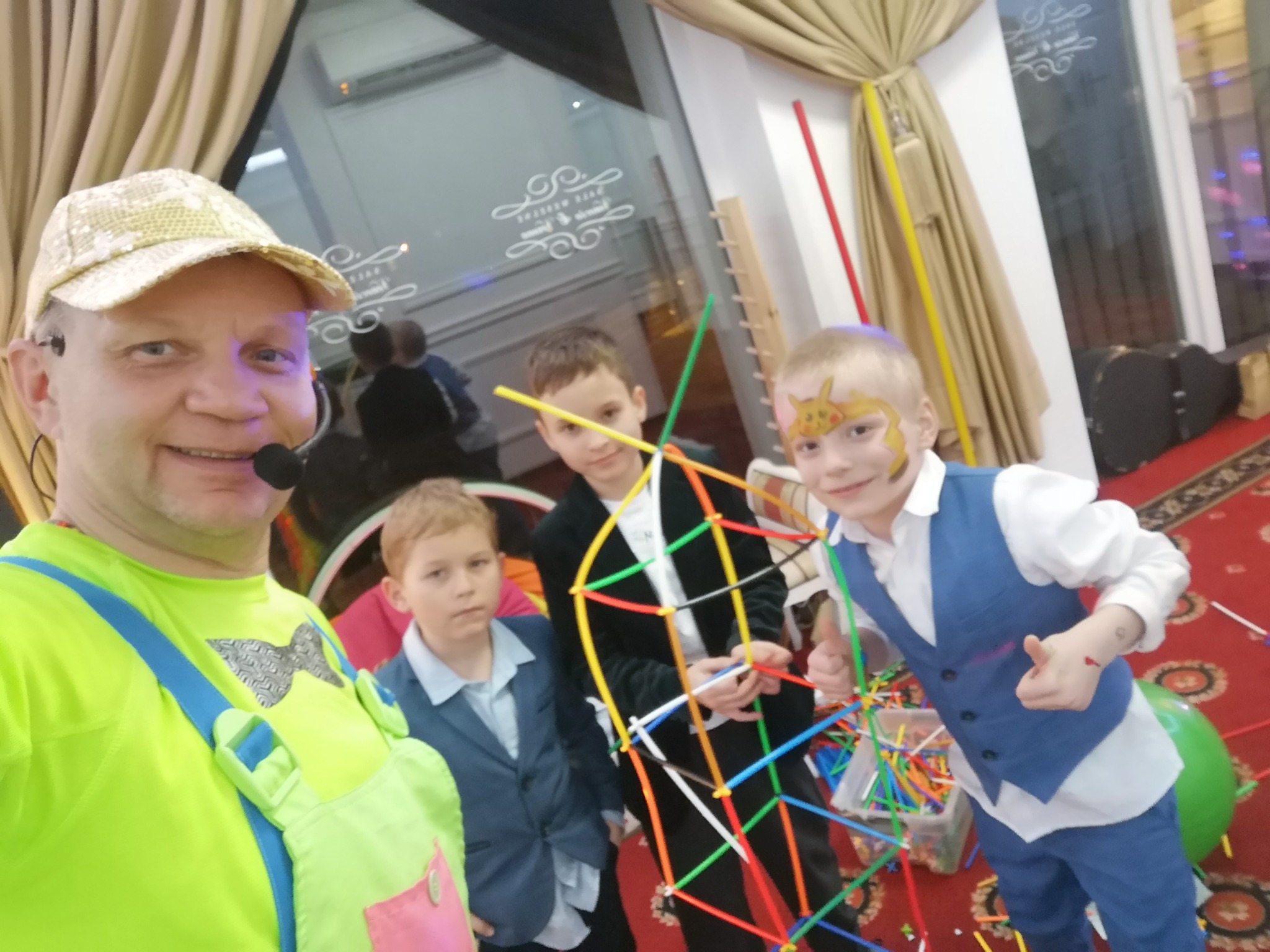 Uśmiechnięty mężczyzna z mikrofonem w neonowo-zielonym stroju i czapce, otoczony przez trzech chłopców, jeden z pomalowaną twarzą, podczas zabawy konstrukcyjnej z kolorowych rurek i łączników.