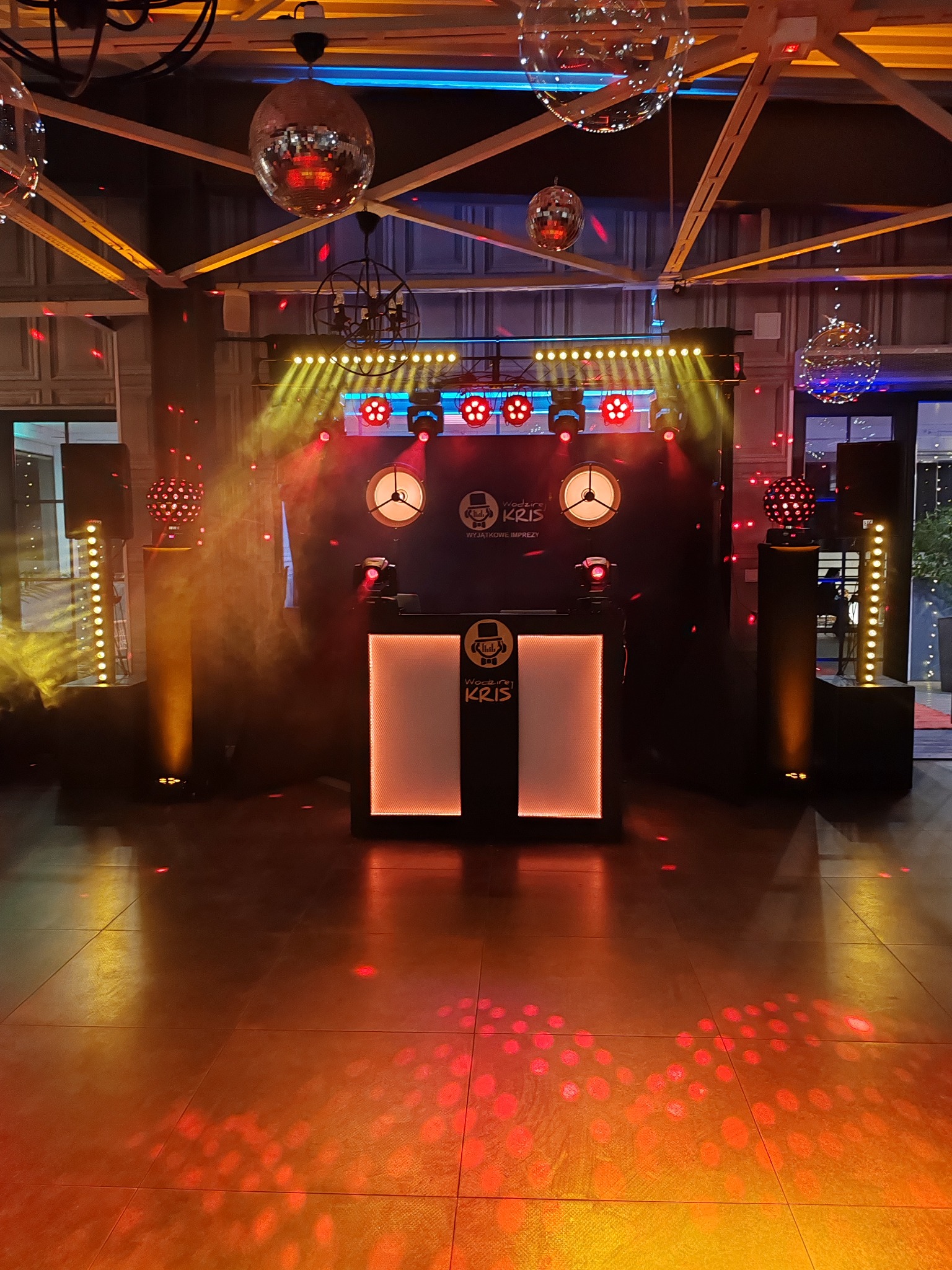 Scena z oświetleniem eventowym: reflektory, kule dyskotekowe, dymiarka. Stanowisko DJ'a z logo. Parkiet oświetlony czerwonymi punktami. Atmosfera imprezy.
