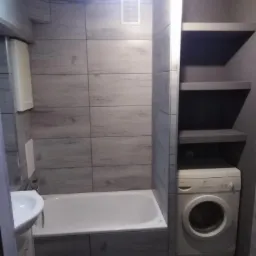 Wykończona łazienka z wanną obudowaną szarymi płytkami imitującymi drewno, umywalką, białą pralką i półkami w zabudowie.