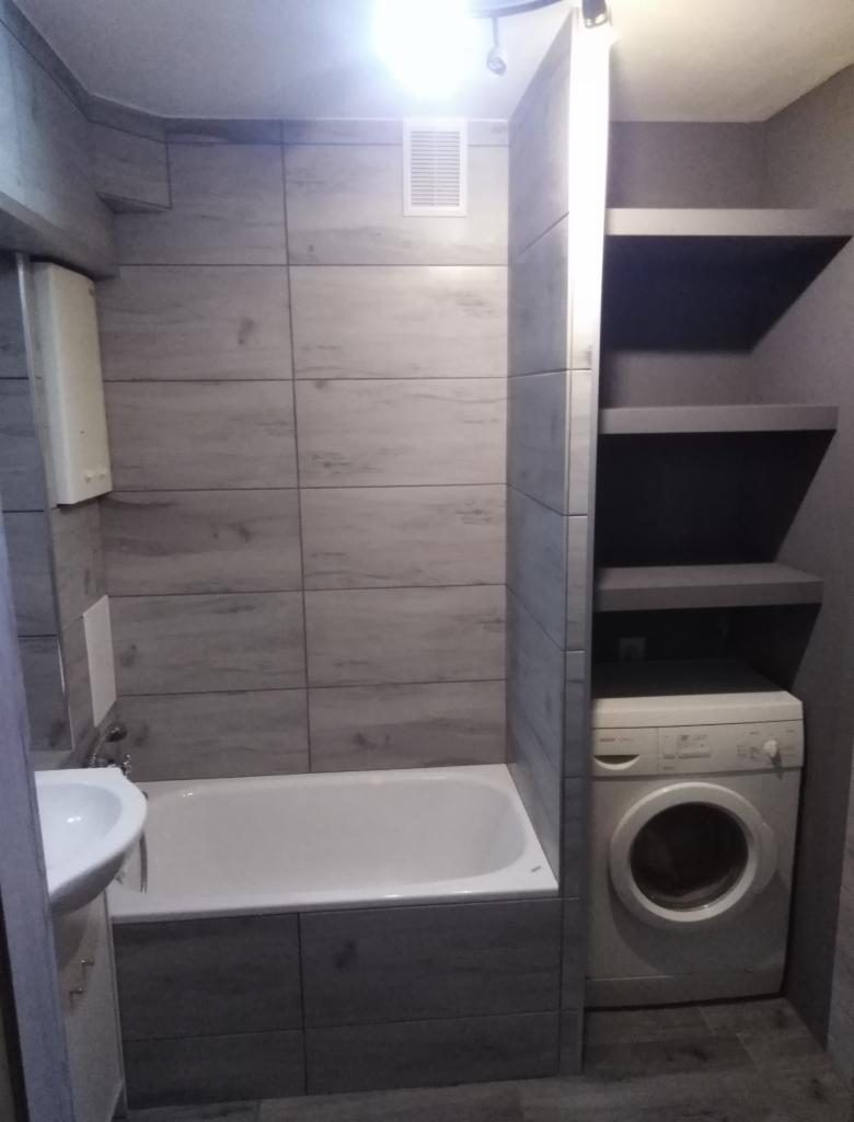 Wykończona łazienka z wanną obudowaną szarymi płytkami imitującymi drewno, umywalką, białą pralką i półkami w zabudowie.