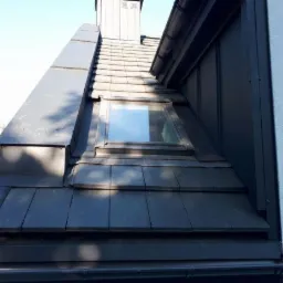 Szczegółowe ujęcie ciemnoszarego dachu z dachówką karpiówką, oknem dachowym i obróbkami blacharskimi, widoczne rynny i fragment elewacji.