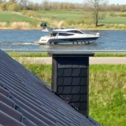 Nowy dach z czarną dachówką ceramiczną i kominem z łupka na tle rzeki z przepływającą motorówką w słoneczny dzień.