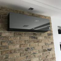 Czarny klimatyzator ścienny o lustrzanej powierzchni zamontowany na ścianie z cegły dekoracyjnej w jasnym odcieniu. Widoczna część sufitu z wbudowanym oświetleniem punktowym.