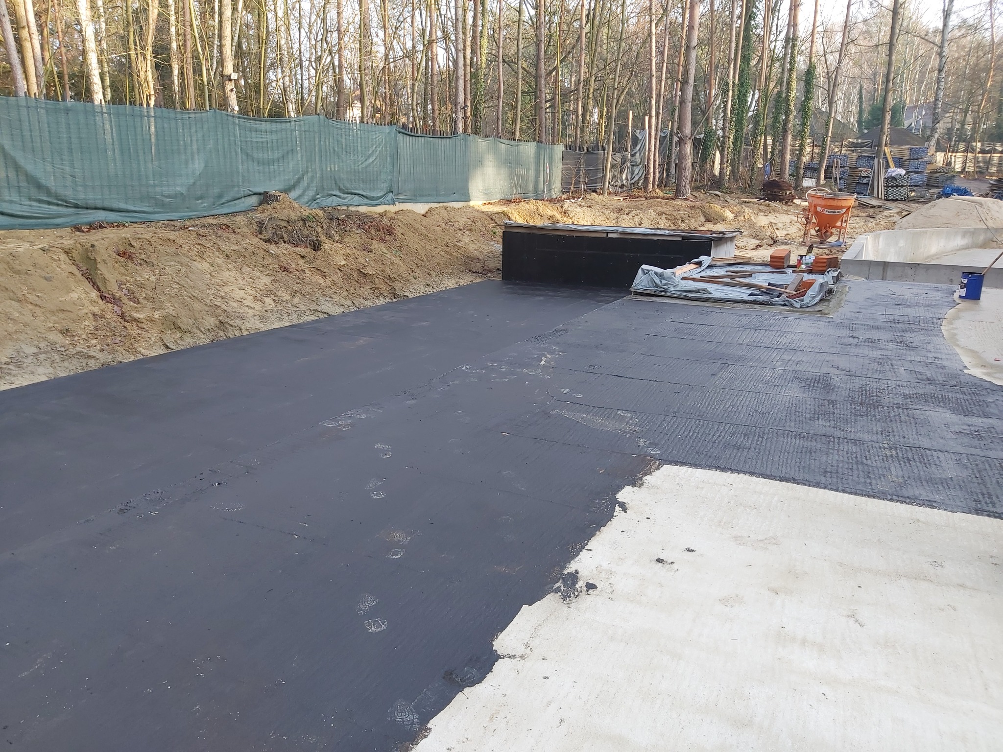 Izolacja fundamentów w trakcie realizacji: widoczna czarna papa podkładowa na izolowanym obszarze, częściowo pokrytym termoizolacją XPS, z folią kubełkową w tle i śladami butów na powierzchni...