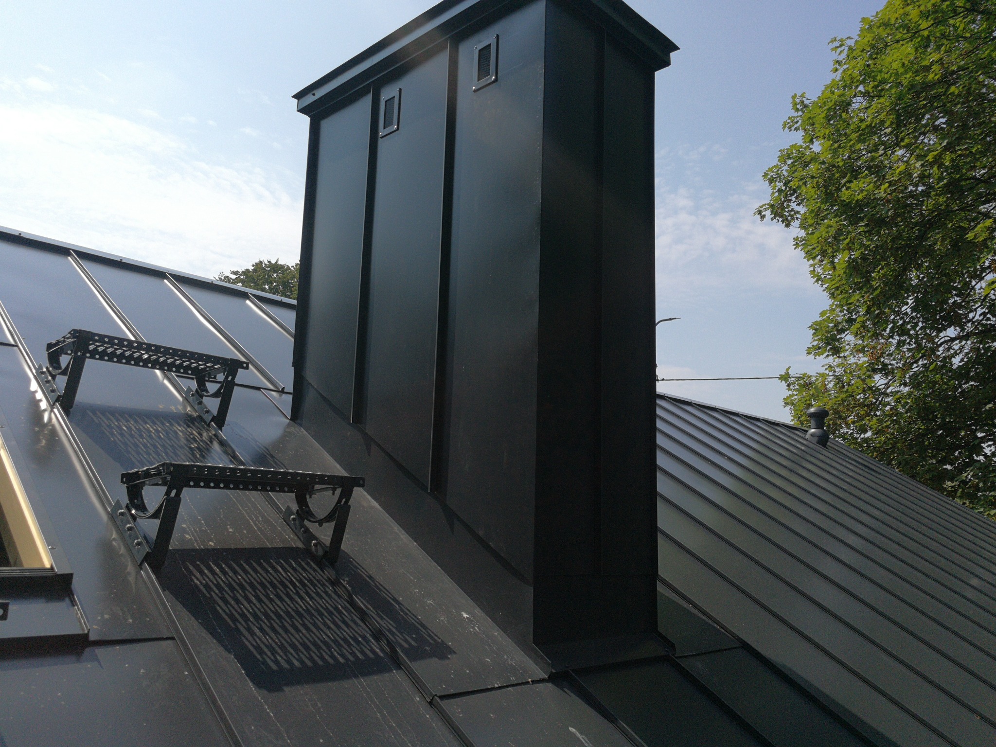 Nowoczesny, czarny komin wentylacyjny na dachu z blachy na rąbek stojący, z zamontowanymi czarnymi stopniami kominiarskimi, na tle błękitnego nieba i zieleni drzew.