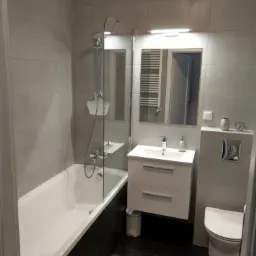 Szara łazienka z wanną za szklanym parawanem, białą szafką z umywalką, wiszącą toaletą i dużym lustrem z oświetleniem.